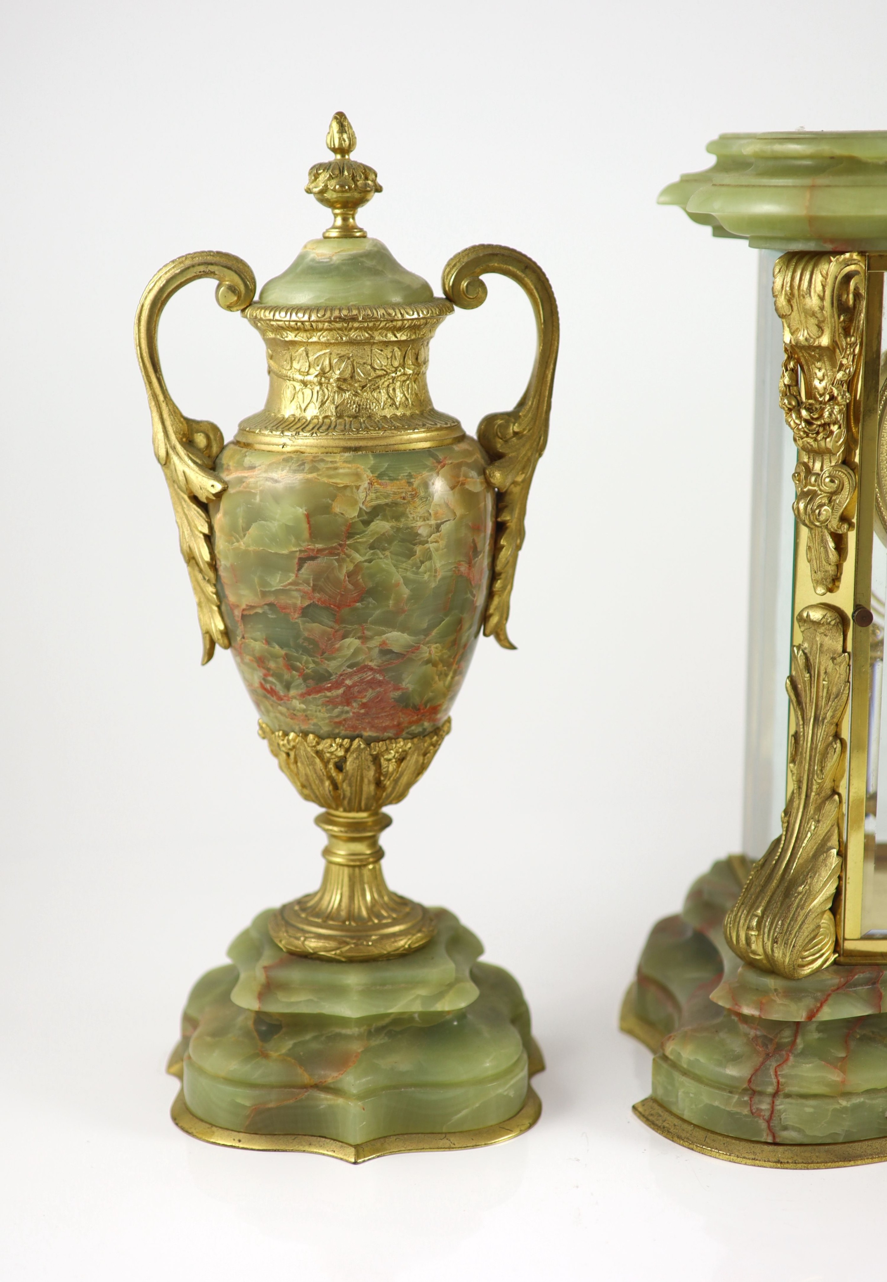 A green onyx and gilt metal clock garniture, comprising a four-glass regulator with enamelled dial in serpentine case and a pair of pedestal urns and covers Clock H 35cm. W 27cm. Urns H 36cm.